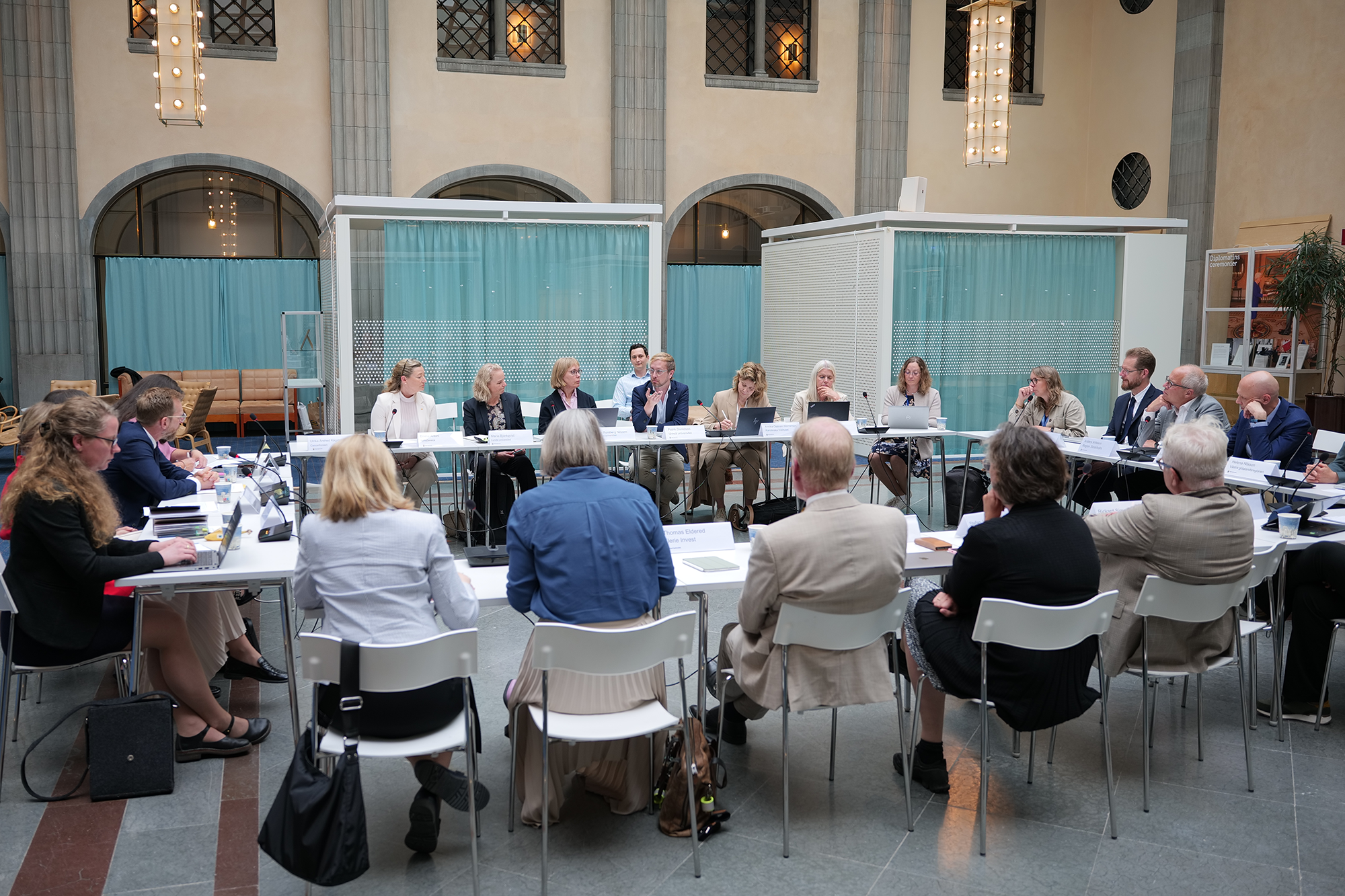Ett tjugotal personer sitter längs fyra långbord som är vända mot varandra. Mötet hålls i en gammaldags banksal med valv och höga fönster.