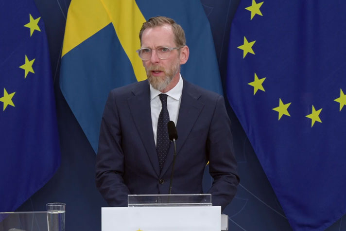Statsrådet står vid en talarpulpet med flaggor i fonden