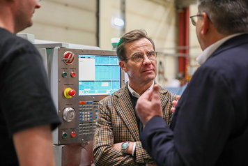 Ulf Kristersson står inomhus framför en maskin och samtalar med två personer.