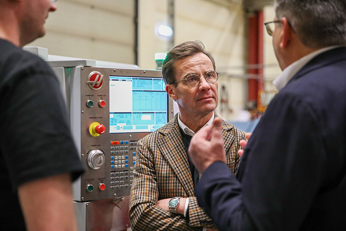 Ulf Kristersson står inomhus framför en maskin och samtalar med två personer.