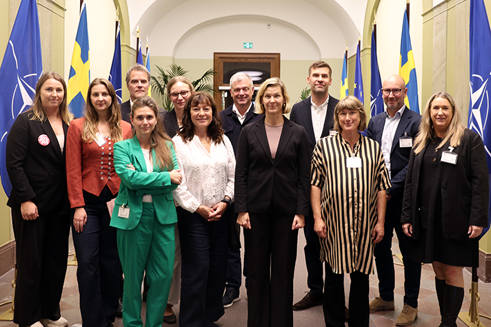 Jämställdhetsministern Nina Larsson står med deltagare från organisationer i en korridor.