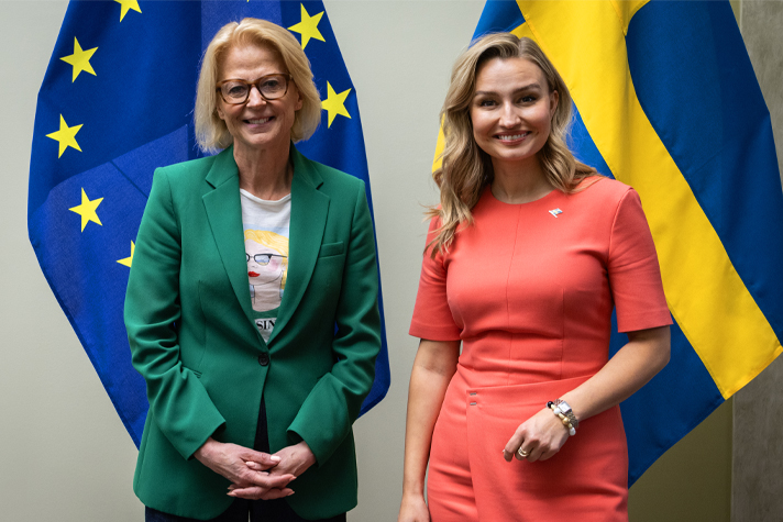 Elisabeth Svantesson och Ebba Busch står sida vid sida och ler mot kameran, framför EU-flaggor och svenska flaggor. Svantesson bär grön kavaj och Busch en korallfärgad klänning.