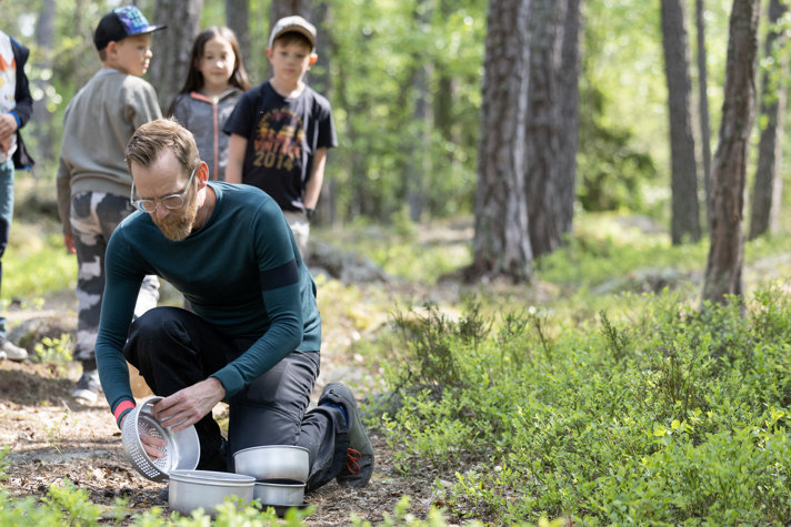 Jakob Forssmed i skogen, sätter ihop ett stormkök. Bakom honom står tre barn.
