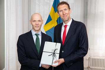 Två män i kostym framför svensk flagga skakar hand och håller i en trycksak. Båda tittar in i kameran.