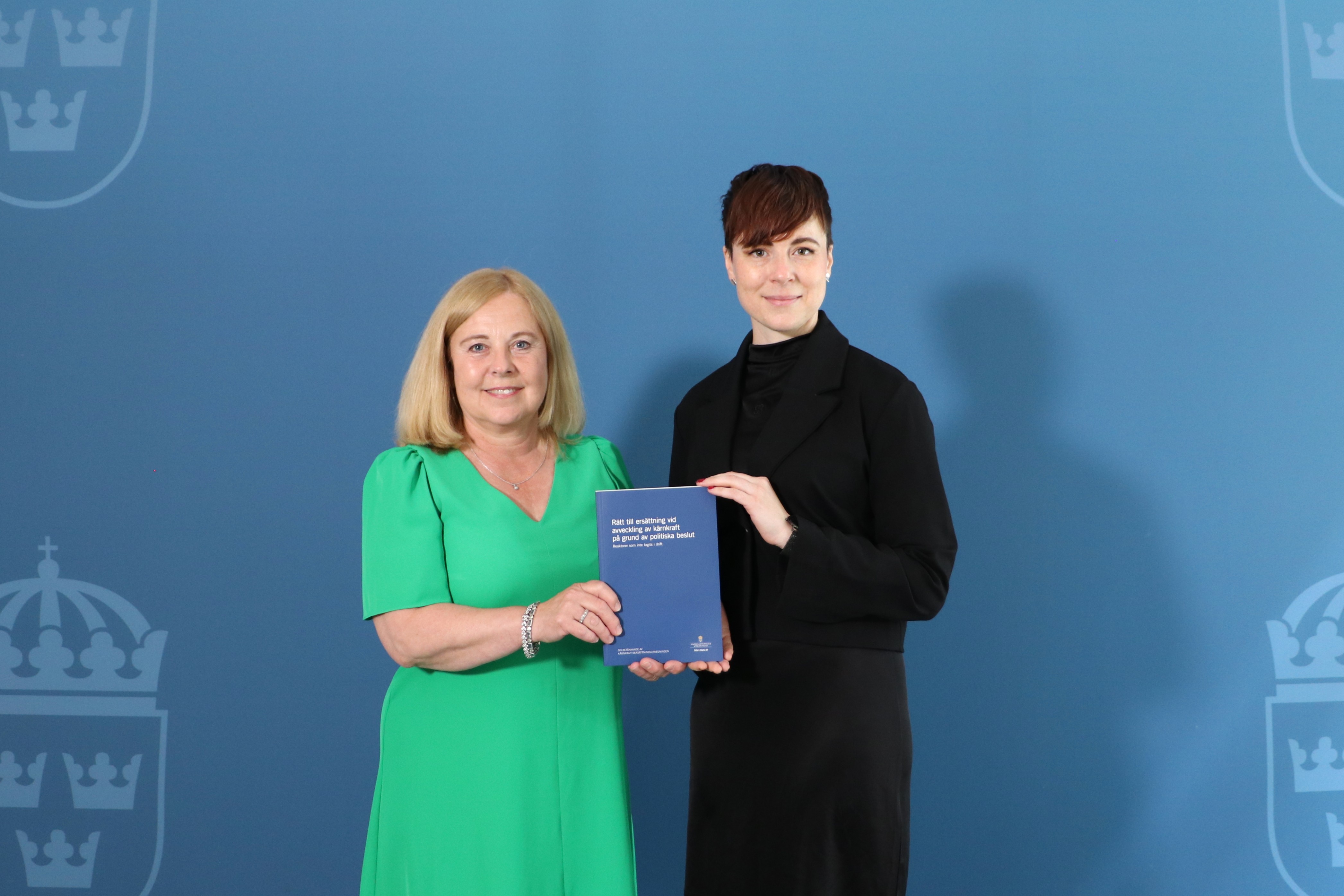 Anne Vadasz Nilsson och Maja Lundbäck står framför en blå bakgrund och håller i ett tryckt exemplar av utredningen