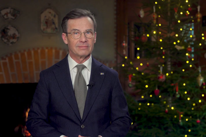 Statsministern är filmad i halvfigur framför en öppen spis samt en julgran i  fonden.