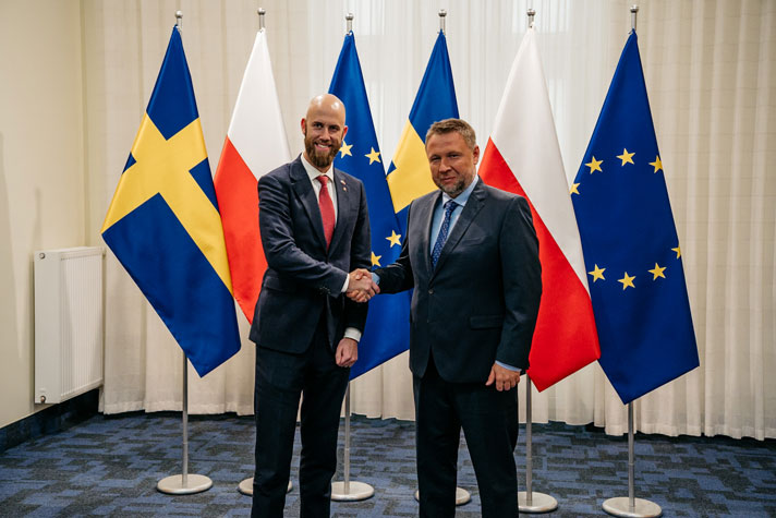 De två ministrarna skakar hand framför flaggor.