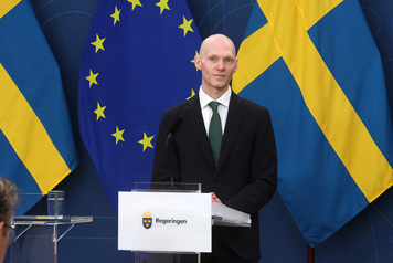 Man i kostym i talarstol med svensk flagga i bakgrunden.