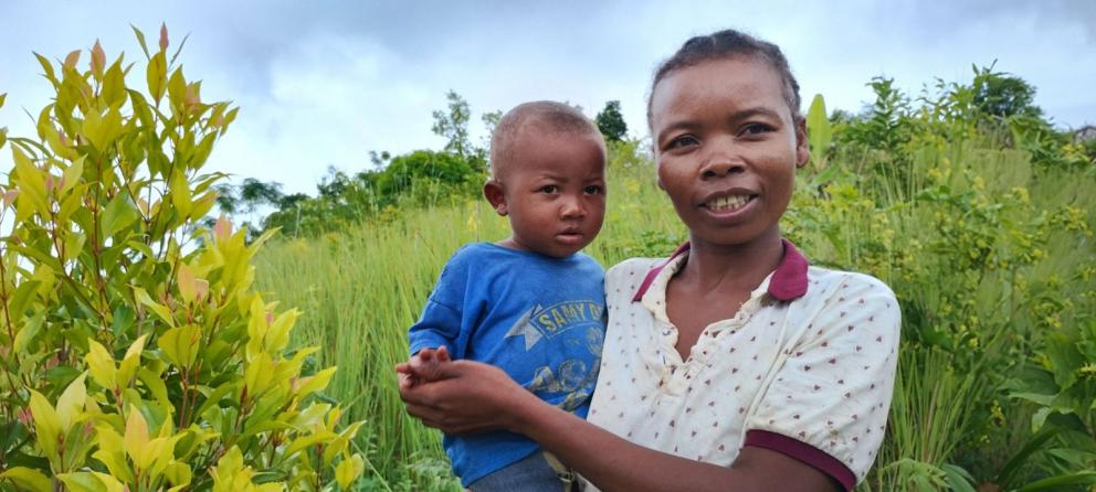 Kvinna med barn i famnen framför växtlighet.