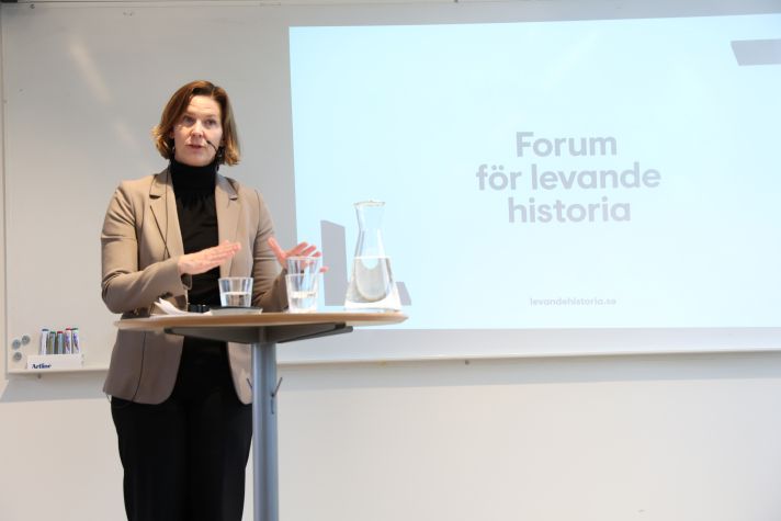 Jämställdhetsministern står vid ett ståbord med glas och vattenkaraff på. I bakgrunden syns en whiteboard med texten Forum för levande historia.