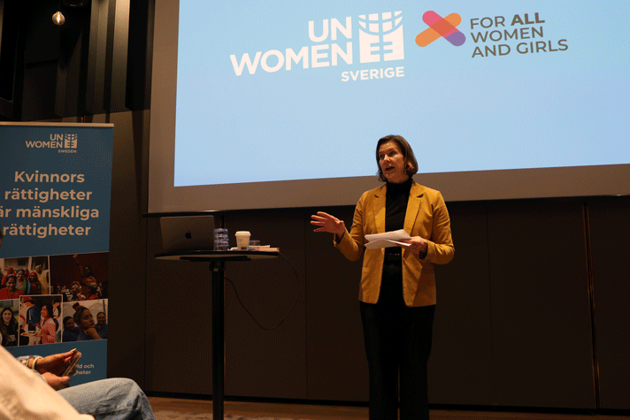 Nina Larsson står framför en skärm med texten "UN Women Sverige- for all women and girls".  Till vänster står en roll-up med texten " Kvinnors rättigheter för mänskliga rättigheter"
