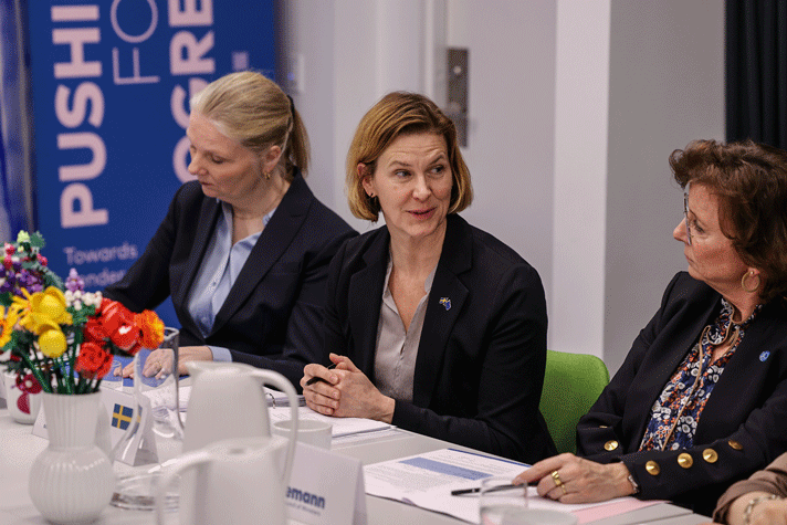 Jämställdhetsminister Nina Larsson sitter på en grön stol vid ett mötesbord och talar. Framför henne på bordet syns mötespapper, en vas med blommor, en vattenkaraff och termosar. I bakgrunden syns en roll-up med texten: Pushing for Progress.