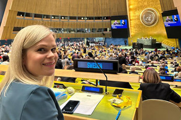 Paulina Brandberg sitter på Sveriges plats i FN:s generalförsamling. Hon tittar in i kameran och ler. I bakgrunden syns många människor som samlats i lokalen.