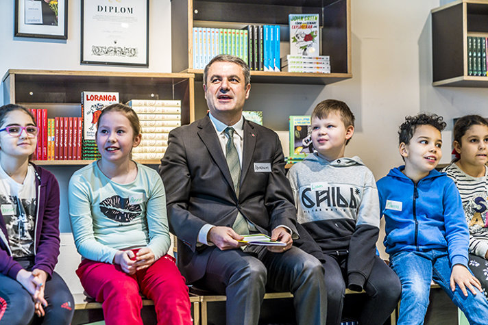 Ministern och flera barn tittar på en skärm. I bakgrunden syns böcker.