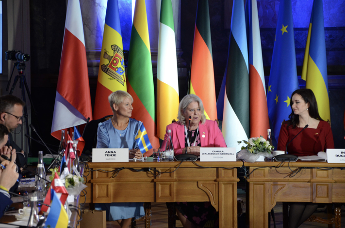 Ministrarna Anna Tenje och Camilla Waltersson Grönvall vid ett mötesbort med flaggor i bakgrunden. På bordet står namnlappar och mikrofoner. 