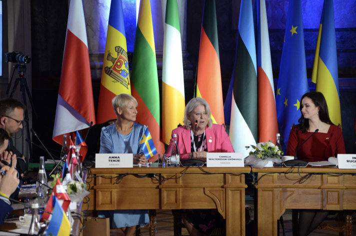 Ministrarna Anna Tenje och Camilla Waltersson Grönvall vid ett mötesbort med flaggor i bakgrunden. På bordet står namnlappar och mikrofoner.