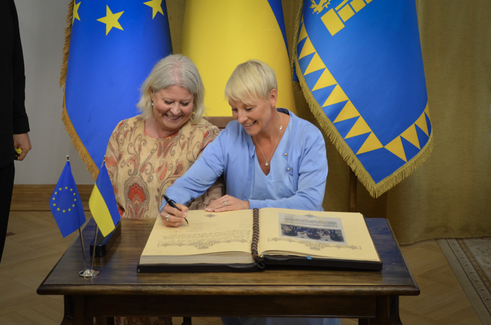 Camilla Waltersson Grönvall och Anna Tenje skriver under i en bok. Bakom dem står ukrainska flaggor i blått och gult. 