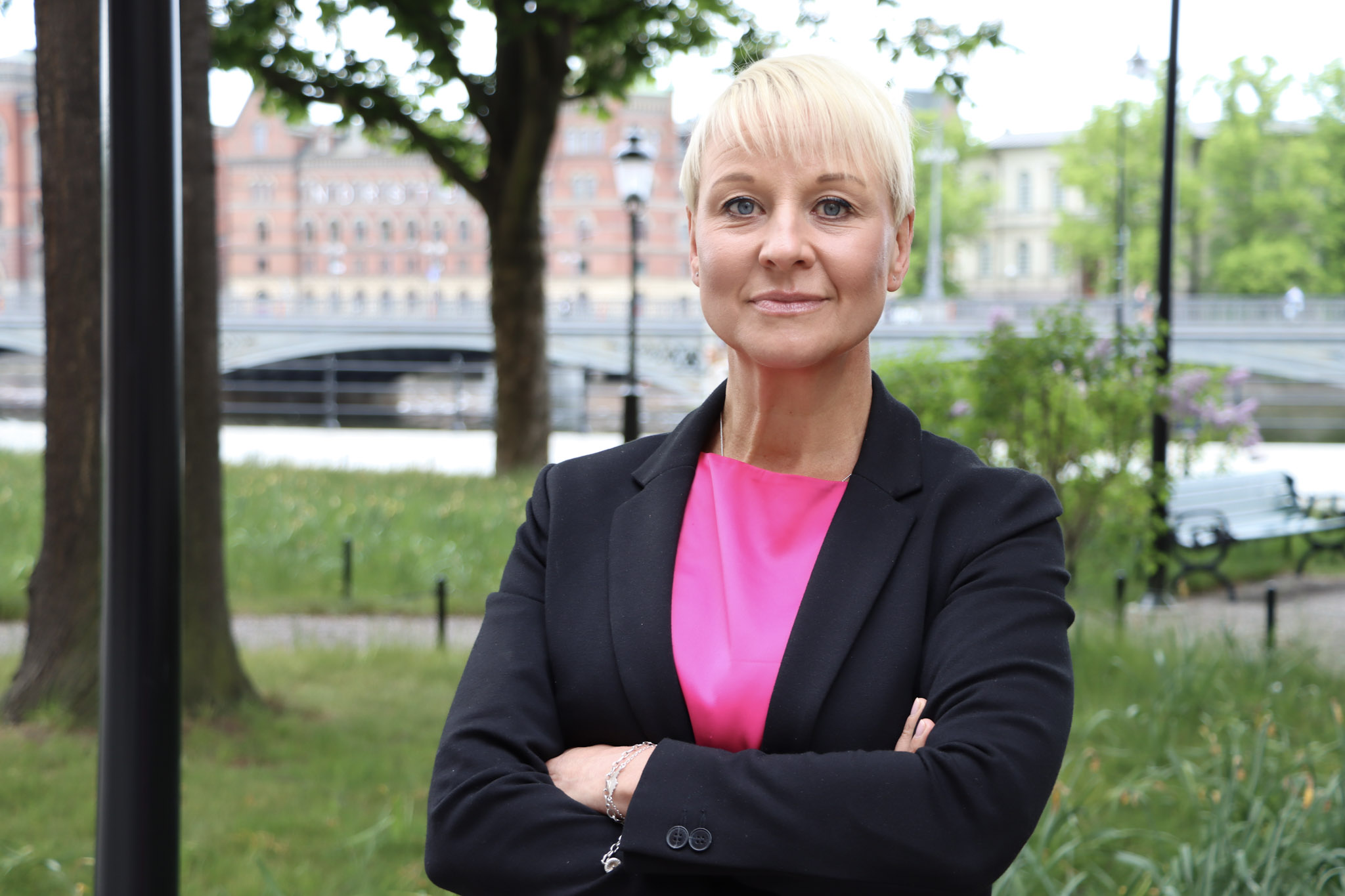 Anna Tenje står vänd mot kameran i parkmiljö, i cerise klänning och mörkblå kavaj.