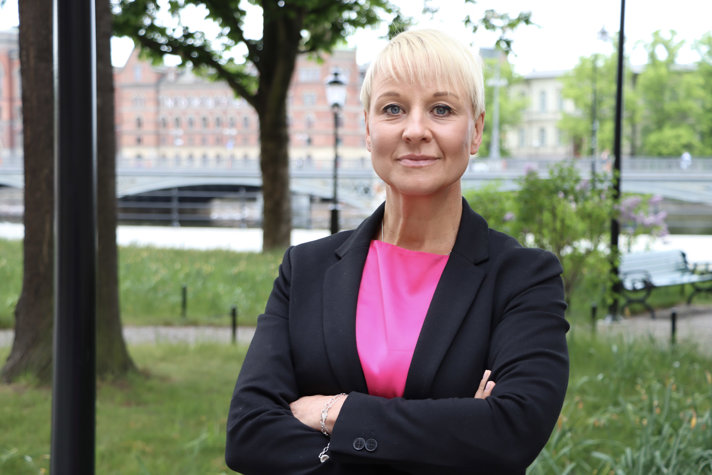 Anna Tenje står vänd mot kameran i parkmiljö, i cerise klänning och mörkblå kavaj.