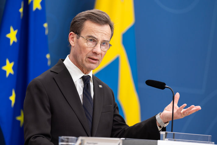 Statsministern står i en talarstol och pratar i en mikrofon. I bakgrunden syns en svenska och en EU-flagga.