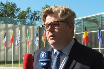 Justitieminister Gunnar Strömmer blir intervjuad av EU-report. Han står utomhus och i bakgrunden syns flera EU-flaggor. 