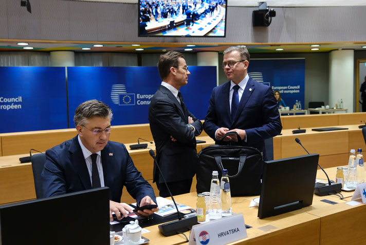 Sverige och Finlands statsministrar står och samtalar i rådets mötesrum bakom sina platser. till vänster sitter Ungers representant och i bakgrunden syns blå skärmar och fler bänkar.