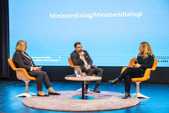 EU-ministern sitter i en gul fåtölj vid ett lågt runt bord och får frågor från Finlands EU-minister. I bakgrunden syns en stor ljusblå skärm med orden Ministerdialog..