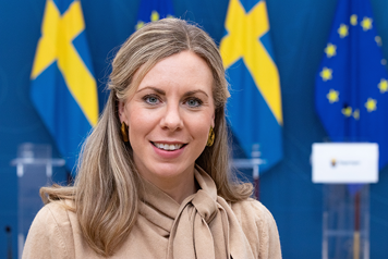 Porträttbild på Jessica Rosencrantz tagen i regeringens pressrum. I bakgrunden syns svenska flaggor och EU-flaggor.