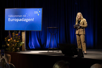 Jessica Rosencrantz talar på en scen inomhus. Till höger om henne på scenen står en skärm med texten "Välkommen till Europadagen. Sverige i EU 30 år.". I förgrunden anas en sittande publik.