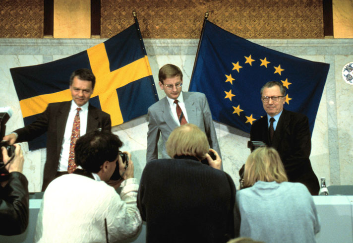 Ulf Dinkelspiel, Carl Bildt och Jaques Delors står framför ett bord. I förgrunden syns några fotografer som tar bilder och i bakgrunden syns svenska flaggan och en EU-flagga.