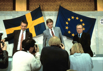 Ulf Dinkelspiel, Carl Bildt och Jaques Delors står framför ett bord. I förgrunden syns några fotografer som tar bilder och i bakgrunden syns svenska flaggan och en EU-flagga.