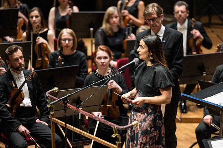 Parisa Liljestrand står vid en mikrofon i Konserthuset. I bakgrunden syns en orkester. 