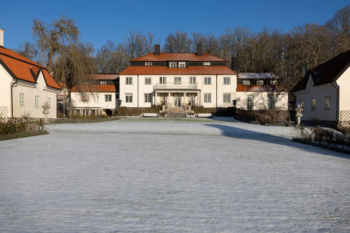 tre vita byggnader med rött tak framför en gårdsplan. Himlen är blå.