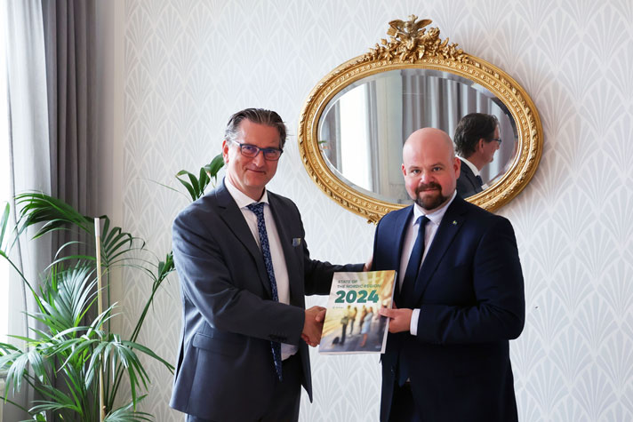 Landsbygdsministern står tillsammans med direktören för Nordregio och tar emot en rapport. I bakgrunden syns en spegel på väggen med guldram.