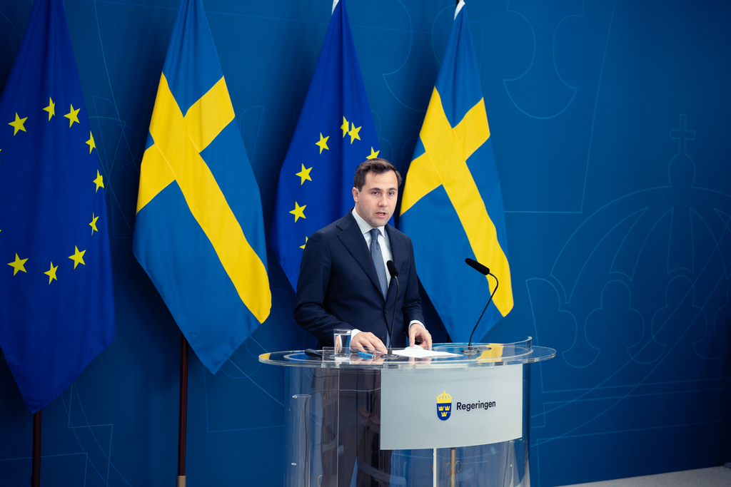Benjamin Dousa står vid talarstol inför svenska flaggor och EU-flaggor.